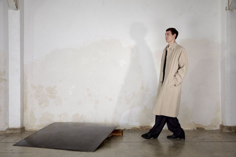 Male model wearing a beige trench coat standing against a weathered white wall with a large cast shadow