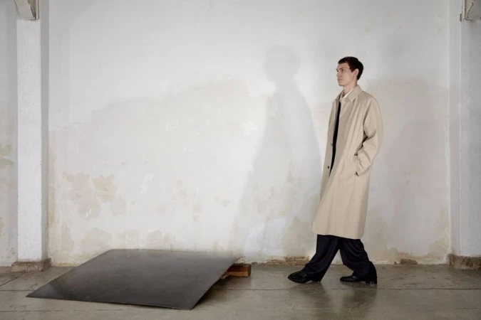 Male model wearing a beige trench coat standing against a weathered white wall with a large cast shadow