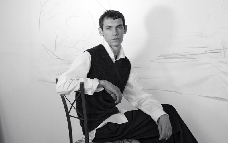 Aleksandr Gordeev seated on a chair in a white studio wearing a white shirt and dark vest for Harper's Bazaar France Homme.