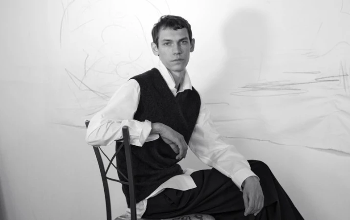 Aleksandr Gordeev seated on a chair in a white studio wearing a white shirt and dark vest for Harper's Bazaar France Homme.