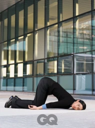 Model lying face down on pavement in a black long-sleeve top and trousers against a glass building exterior for GQ Korea.