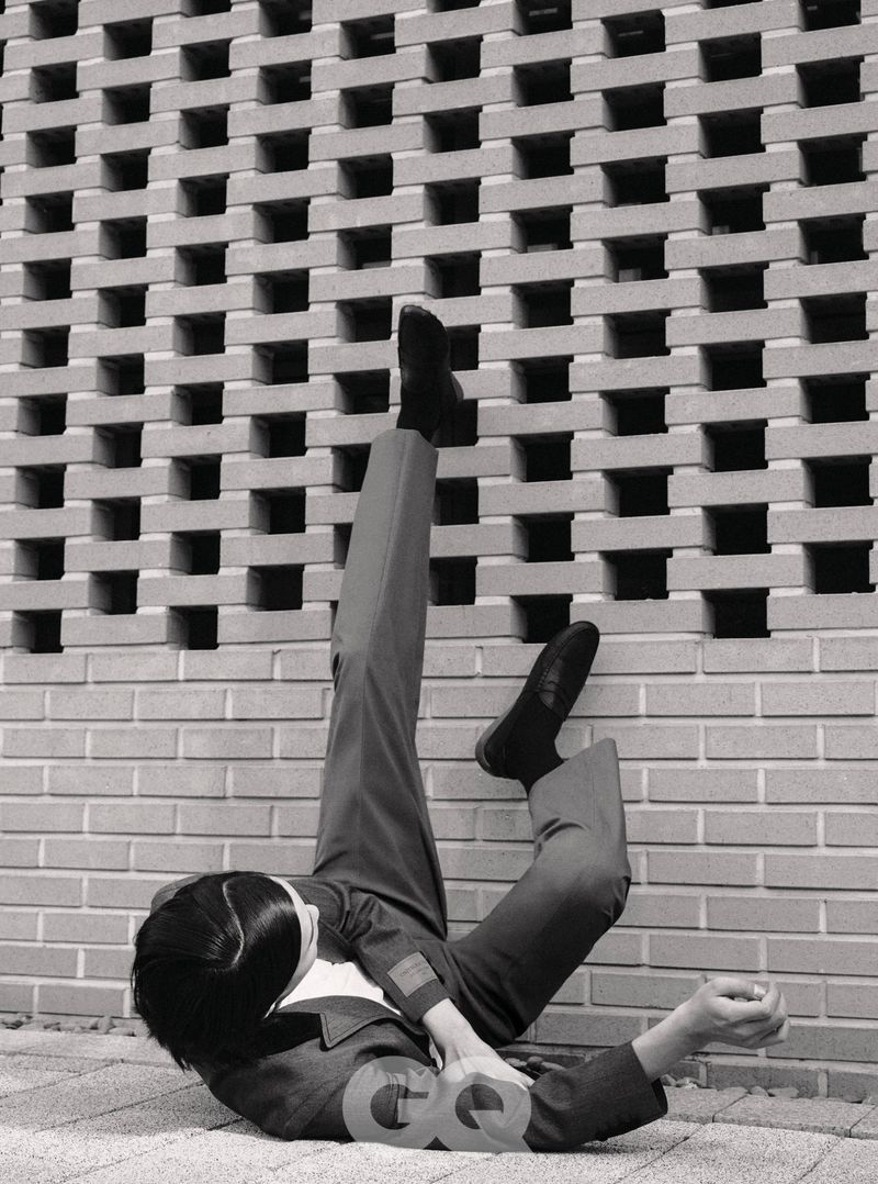 Model lying on the ground with legs kicked up against a textured concrete breeze block wall in GQ Korea editorial.