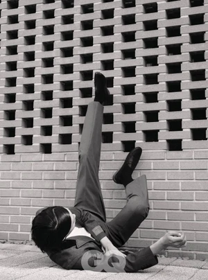 Model lying on the ground with legs kicked up against a textured concrete breeze block wall in GQ Korea editorial.