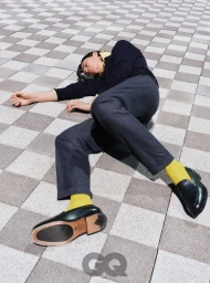 Model lying on a patterned checkered tile floor wearing dark trousers, a navy sweater, yellow socks, and leather loafers