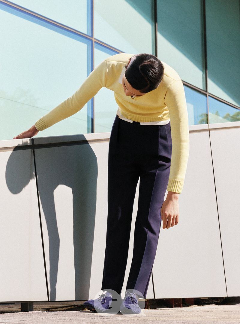 Model wearing a pale yellow sweater and dark trousers leaning forward against a modern architectural exterior wall.