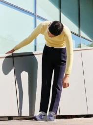 Model wearing a pale yellow sweater and dark trousers leaning forward against a modern architectural exterior wall.
