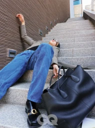 Model lying backward on outdoor stone stairs wearing blue jeans, a grey jacket, and black leather loafers with a large black leather tote bag.