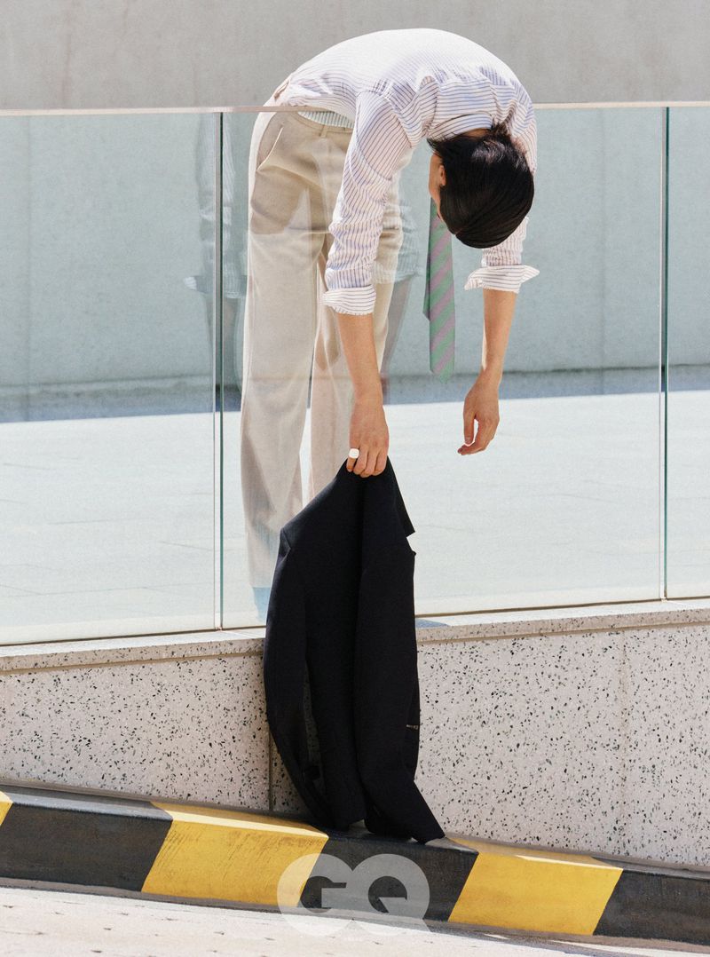 Model bending forward over a glass balcony railing while holding a dark jacket for GQ Korea