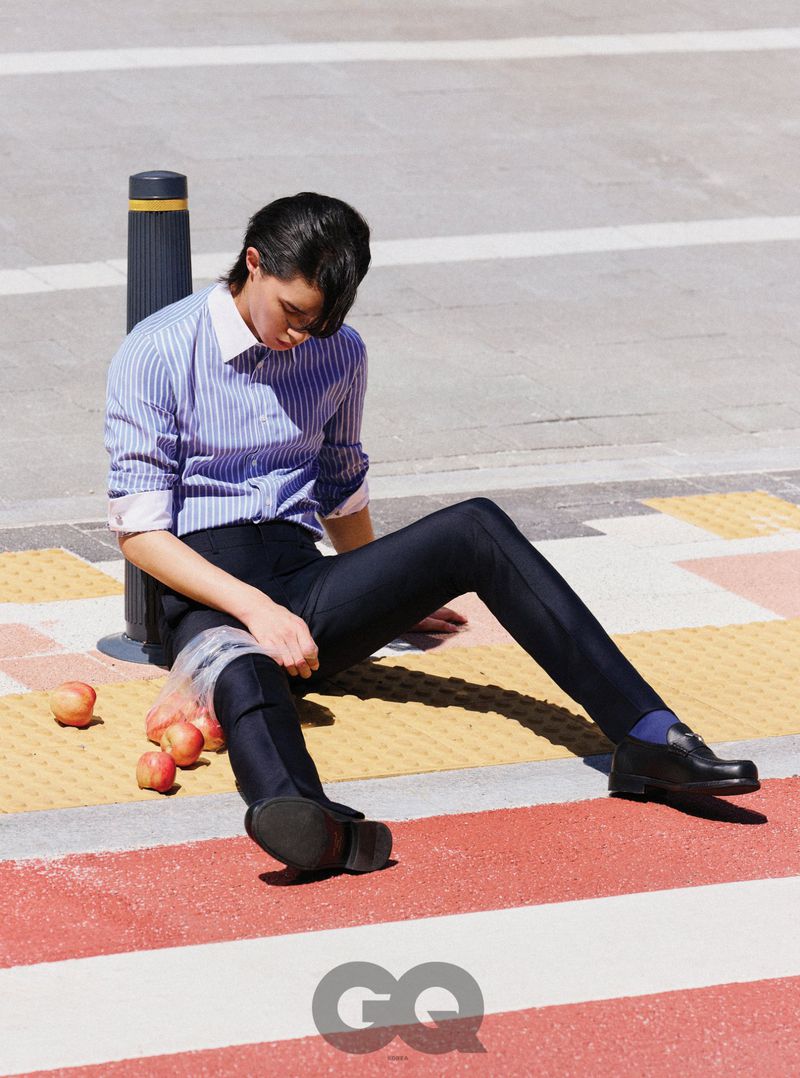 Model seated on a city street next to a plastic bag of apples wearing a blue striped shirt and dark trousers for GQ Korea
