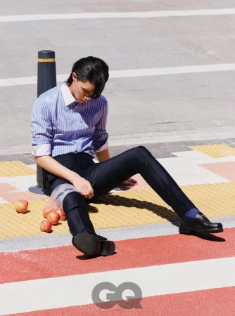Model seated on a city street next to a plastic bag of apples wearing a blue striped shirt and dark trousers for GQ Korea