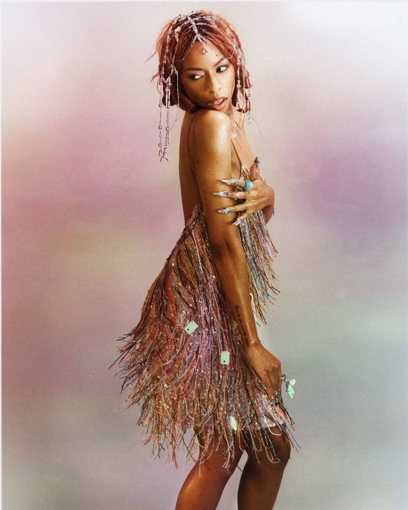 Ravyn Lenae wearing a fringed, shimmering dress with beaded hair accessories against a soft gradient backdrop.