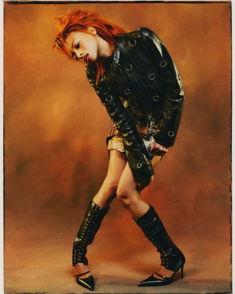 Ravyn Lenae in a black leather jacket and knee-high boots posing against a warm brown gradient backdrop for Teen Vogue.