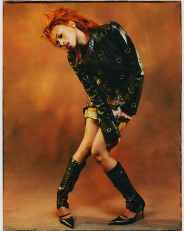 Ravyn Lenae in a black leather jacket and knee-high boots posing against a warm brown gradient backdrop for Teen Vogue.
