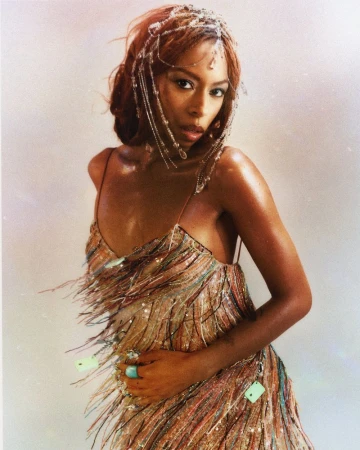 Portrait of singer Ravyn Lenae wearing a fringed, sequined dress and a beaded headpiece against a neutral background.
