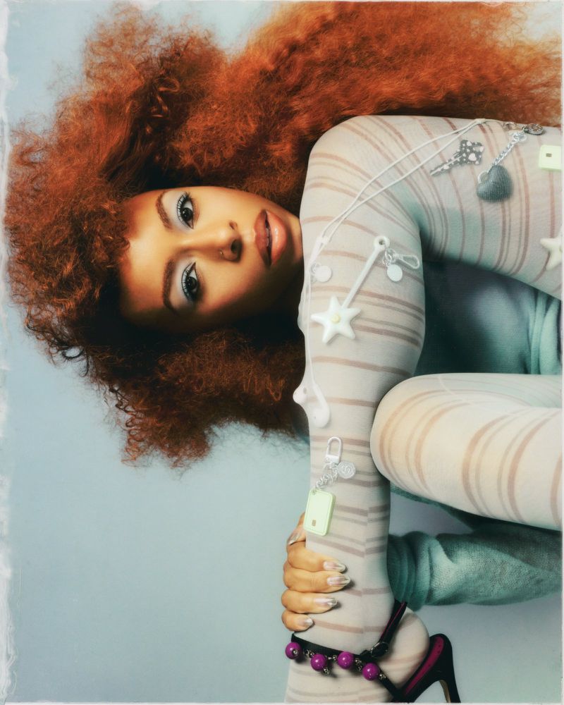 Portrait of Ravyn Lenae with vibrant red hair wearing striped tights against a neutral backdrop for Teen Vogue.