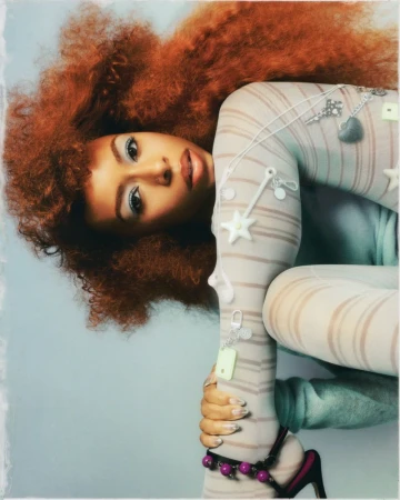 Portrait of Ravyn Lenae with vibrant red hair wearing striped tights against a neutral backdrop for Teen Vogue.