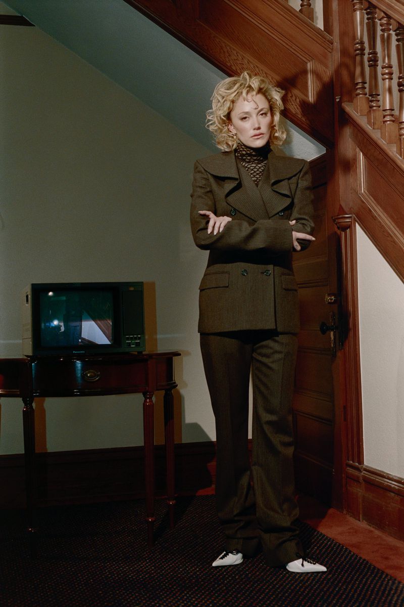 Maika Monroe standing with arms crossed wearing a dark suit in an interior hallway with a television on a side table