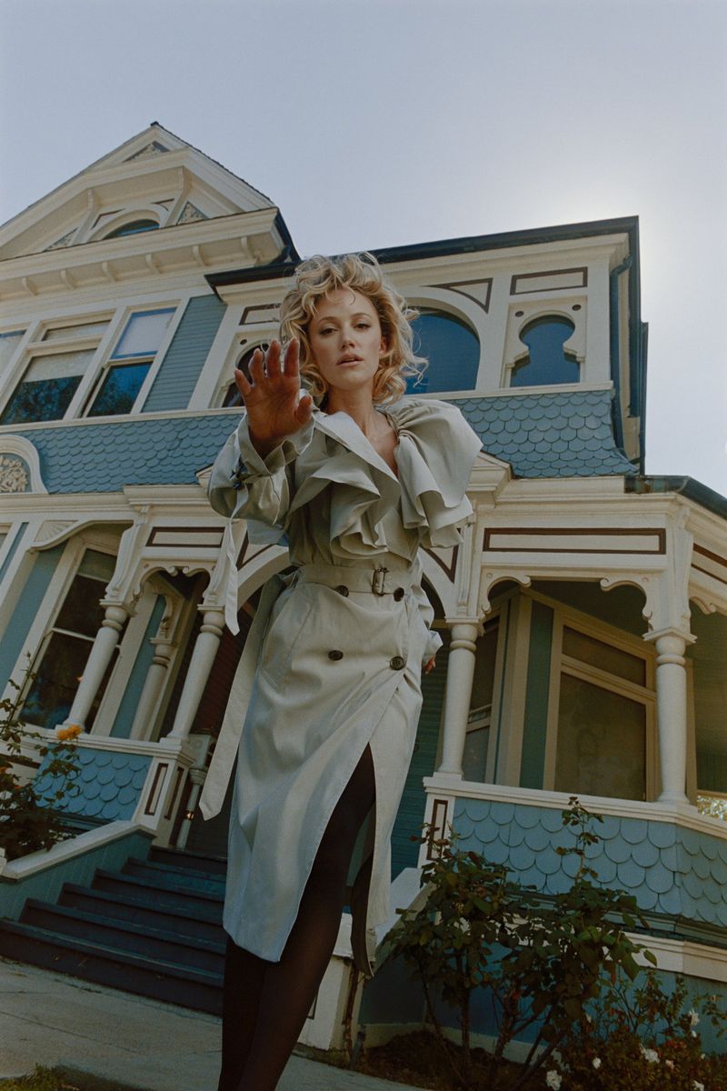Maika Monroe wearing a light grey belted trench coat standing before a large blue and white Victorian house