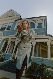 Maika Monroe wearing a light grey belted trench coat standing before a large blue and white Victorian house