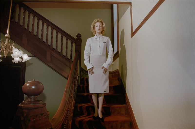 Maika Monroe standing on a wooden staircase wearing a white shirt and beige skirt for Interview Magazine.