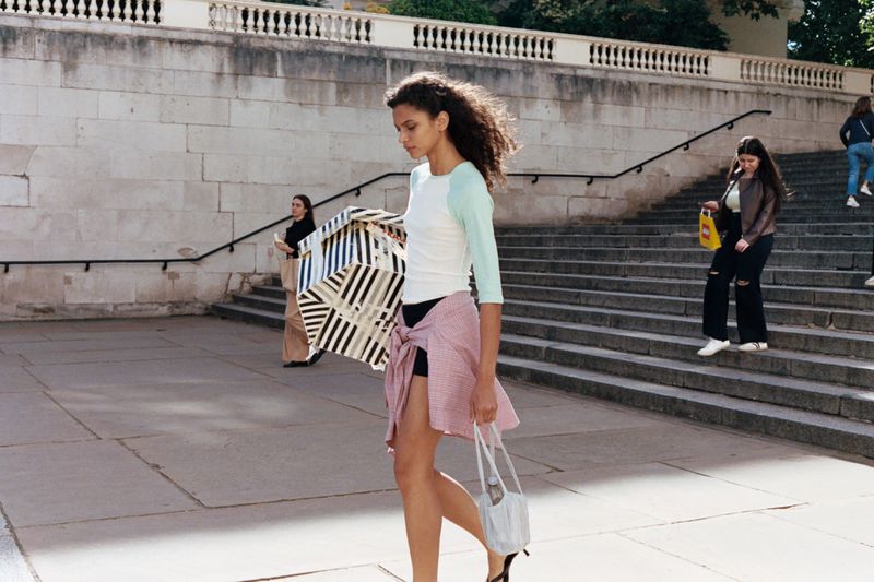Model walking outdoors carrying a striped box and handbag, with stone steps and other people in the background.