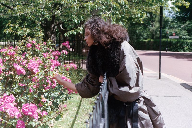 Model with dark curly hair wearing an oversized coat with a dark fur collar touching pink rose flowers in a park.
