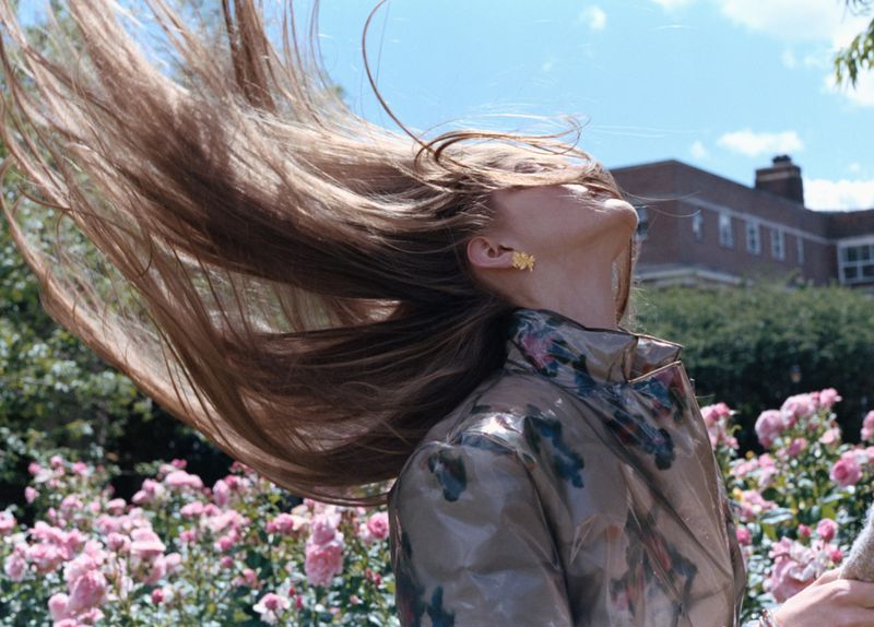 Model flipping long brown hair in motion wearing a patterned jacket outdoors among blooming pink roses.