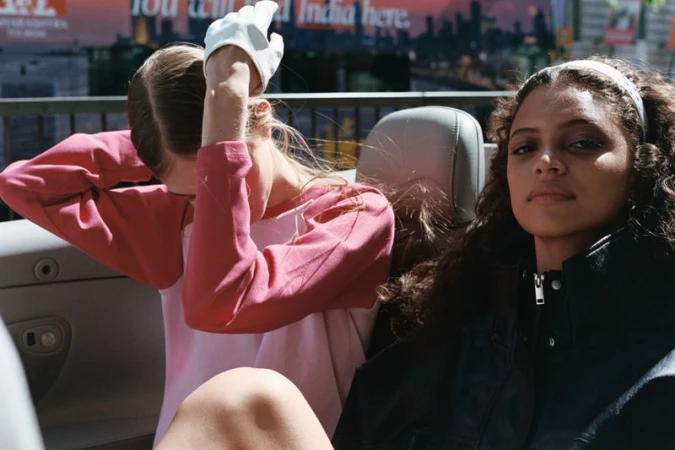 Two models seated inside a convertible car, one adjusting a white glove and the other looking towards the camera.