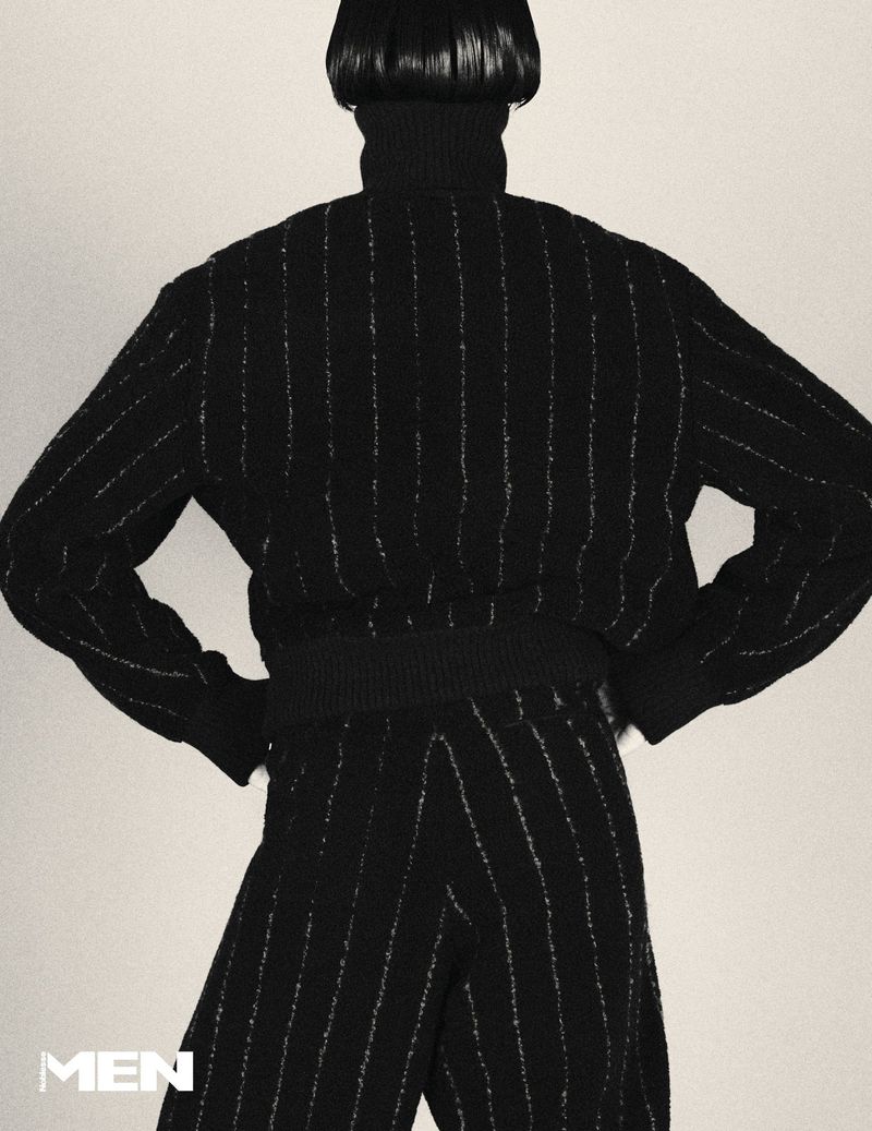Model seen from behind wearing a black pinstripe turtleneck sweater and matching trousers against a pale background