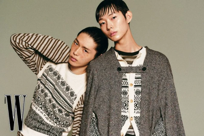 Two male fashion models posing against a grey seamless backdrop wearing patterned knitwear and a grey tweed cape.
