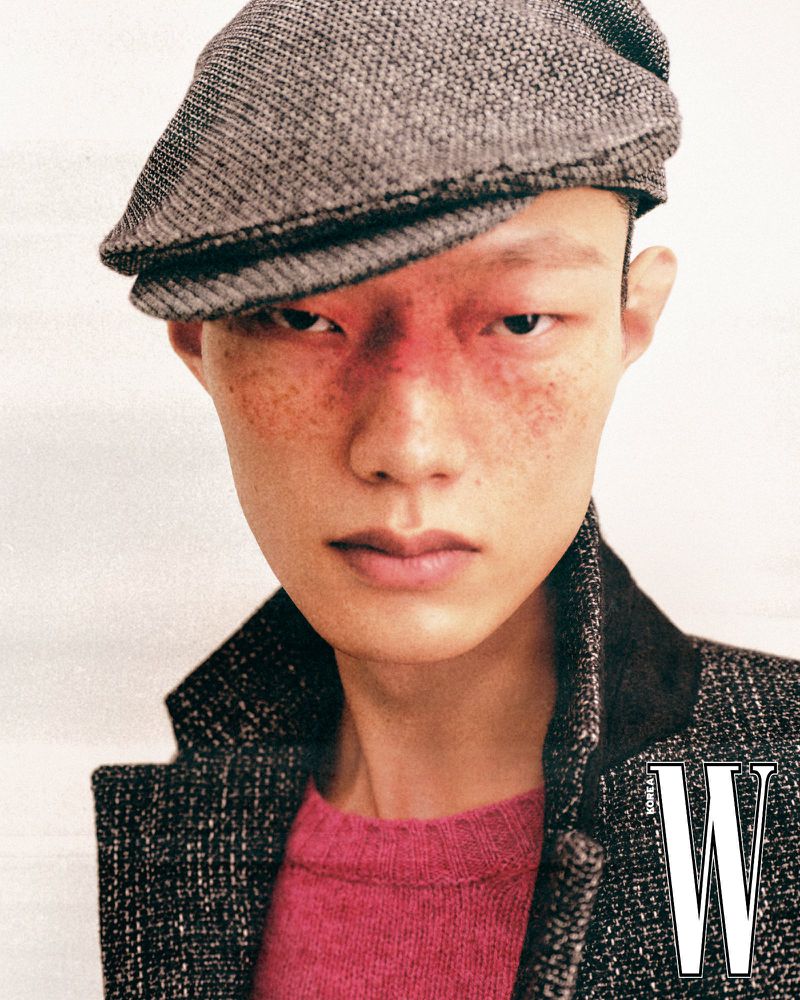 Close-up portrait of an Asian male model wearing a grey flat cap, tweed jacket, and pink sweater against a plain background.