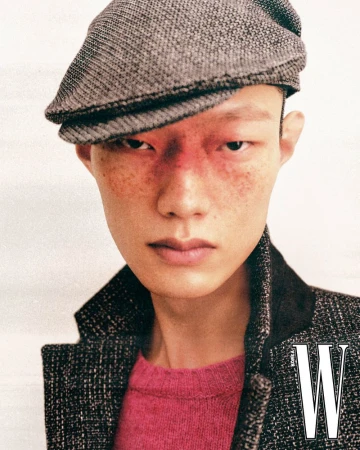Close-up portrait of an Asian male model wearing a grey flat cap, tweed jacket, and pink sweater against a plain background.