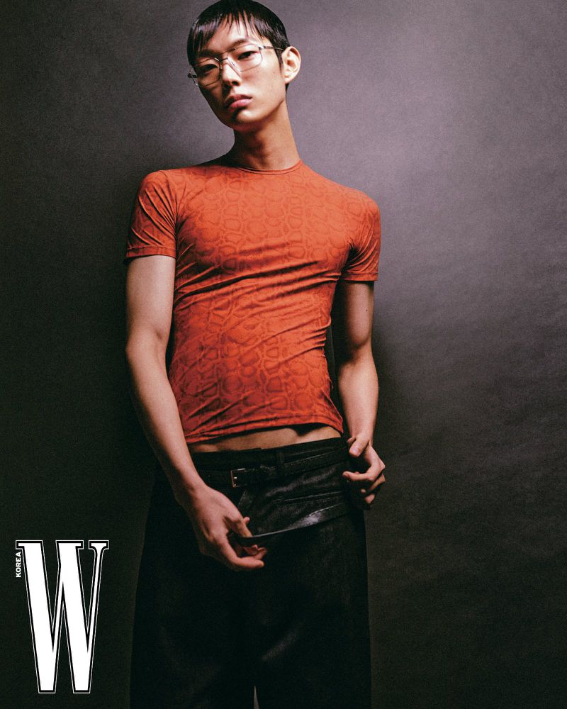 Asian male model wearing a rust orange patterned t-shirt and dark trousers against a textured dark background for W Korea.