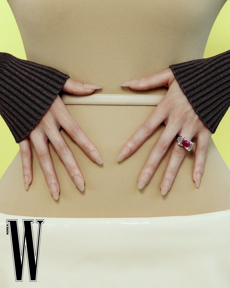 Close-up of hands resting on a waist, wearing brown cuffs and a silver ring with a red stone against a yellow background.