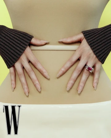 Close-up of hands resting on a waist, wearing brown cuffs and a silver ring with a red stone against a yellow background.