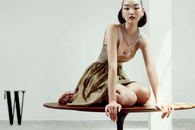 Model kneeling on a dark wooden oval table wearing a beige dress and jewelled necklace against a white wall.