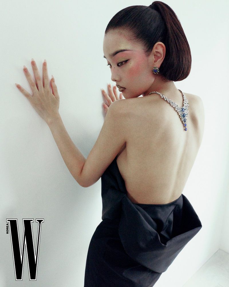 Model Bomi Youn posing against a white wall wearing a backless black dress, diamond necklace, and blue earrings for W Korea by Kim Hee June