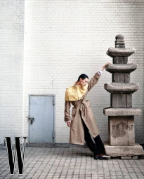 Model Hoyong Kim leaning against a stone pagoda, wearing a brown trench coat with a yellow cape detail.