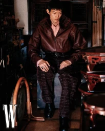 Asian male model wearing a dark leather bomber jacket and patterned trousers seated indoors in W Korea editorial.