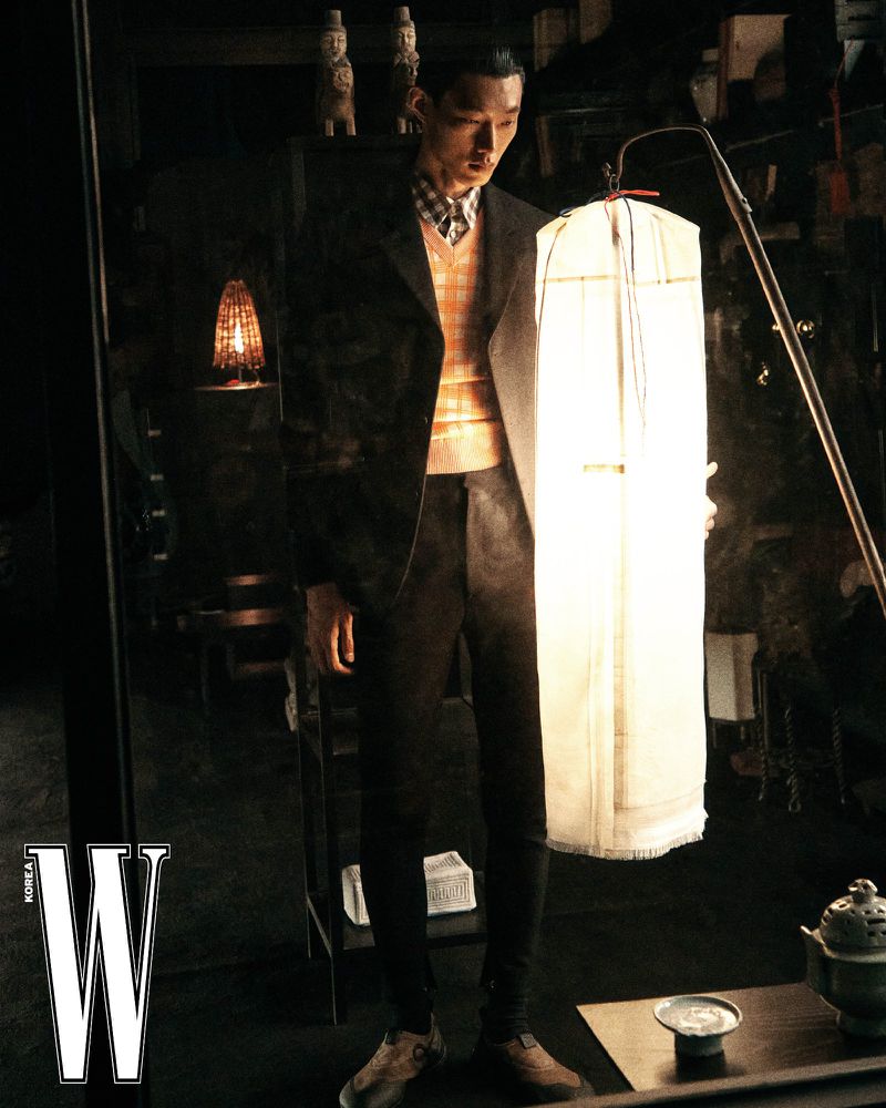 Model Hoyong Kim wearing a dark jacket, patterned vest, and trousers next to an illuminated garment bag for W Korea