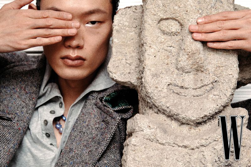 Close-up of a model covering his eyes alongside a carved stone sculpture, wearing a textured tweed blazer and grey polo shirt for W Korea.