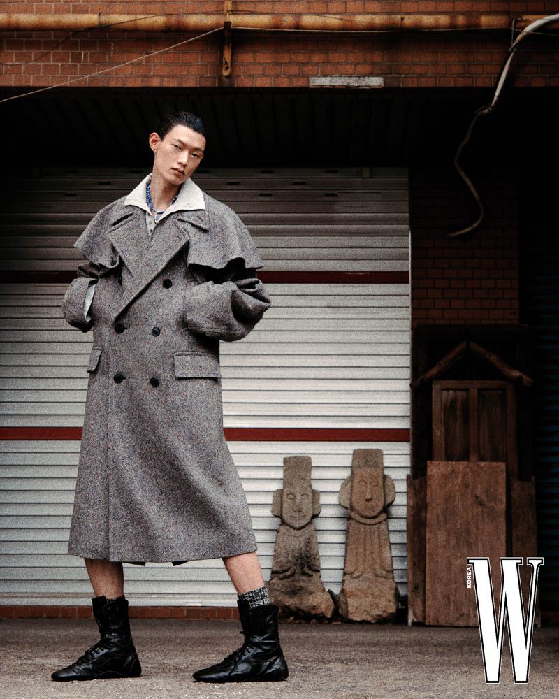 Model Hoyong Kim in a grey double-breasted cape coat and black boots standing outdoors in front of a shutter door.