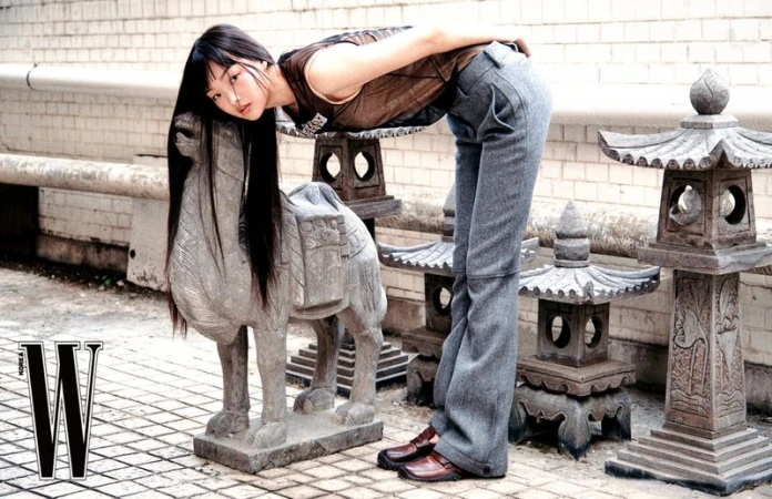 Model with long dark hair bending over an antique stone animal statue next to stone lanterns for W Korea
