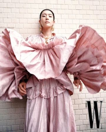 Model in a voluminous pink ruffled dress standing against a light brick wall for W Korea fashion editorial.