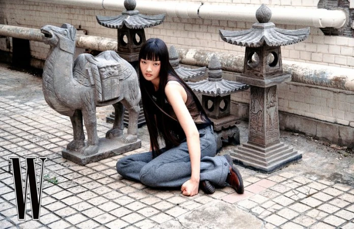 Model with long dark hair kneeling on tiled floor next to stone camel statue and miniature pagoda lanterns.