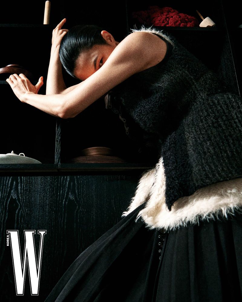 Model wearing a black sleeveless textured top and dark pleated skirt poses with arms raised against a dark shelving unit