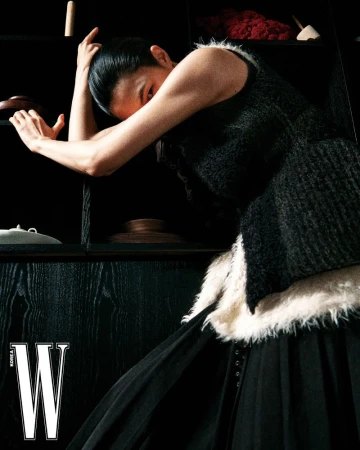 Model wearing a black sleeveless textured top and dark pleated skirt poses with arms raised against a dark shelving unit