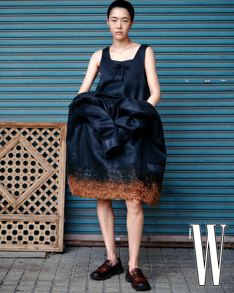 Model wearing dark sleeveless top and jacket standing in front of blue roller shutter door with wooden frame on ground