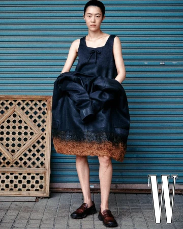 Model wearing dark sleeveless top and jacket standing in front of blue roller shutter door with wooden frame on ground