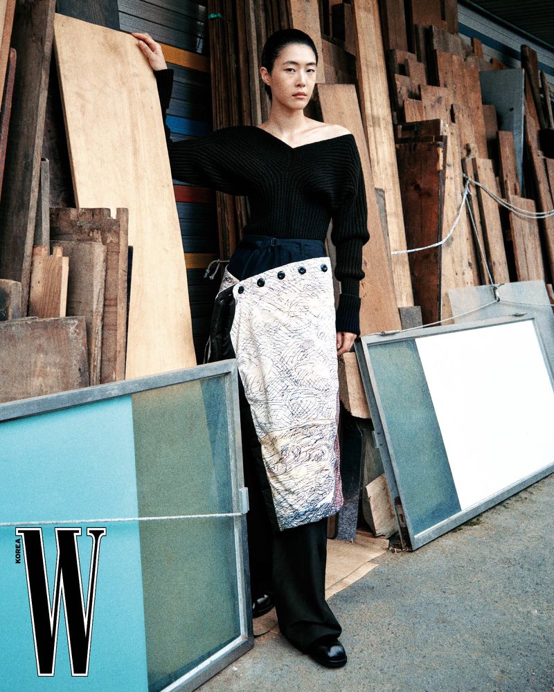 Model wearing a black off-the-shoulder top and patterned wrap skirt in an industrial timber yard